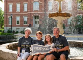 Two kids posing with their grandparents in front of Purdue's John Purdue Fountain holding a Grandparents University sign